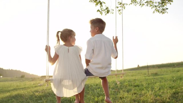 SLOW MOTION CLOSE UP: Couple of happy kids hugging and swinging on wooden swing at golden sunset. Brother embracing little sister on a swing. Young love between boy and girl on romantic summer evening
