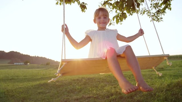 SLOW MOTION CLOSE UP: Smiling little girl swaying on swing at golden summer sunset. Cheerful young girl in white dress swinging over a setting sun in park. Female child enjoying youth playing outside