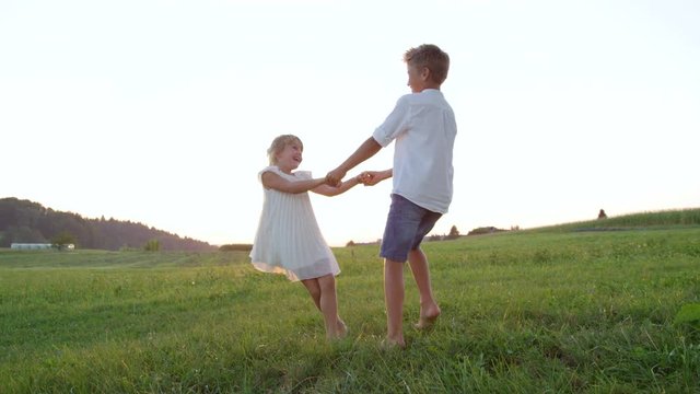 SLOW MOTION LENS FLARE: Cheerful kids holding hands and dancing at golden sunset. Playful brother and sister playing ring around the Rosie in meadow field. Lively boy and girl having fun in summer