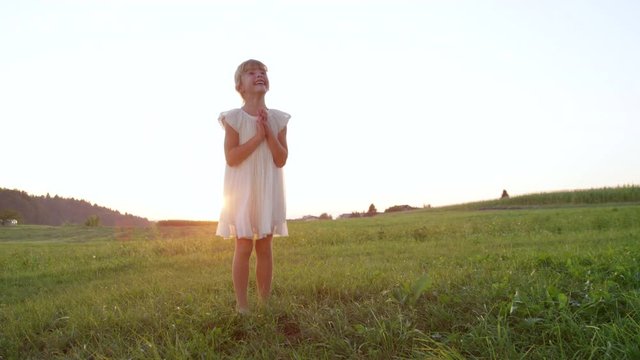 SLOW MOTION CLOSE UP Silly little girl in white dress having fun outdoors in late afternoon. Goofy child enjoying her summer playing in a meadow at sundown. Excited little sister clapping cheerfully.