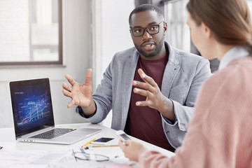 Crew of talented young employees explain ideas during working meeting. Black male employer shows financial presentation, analyze graphics, gestures with hands, look attentively at interloutor