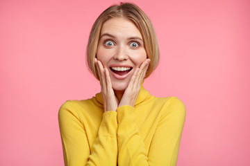 Happy excited female has light hair, glad to see something unexpected, keeps hands on cheeks, smiles broadly, isolated over pink background. Overjoyed woman stares at camera with joyful expression