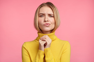 Portrait of beautiful woman curves lips, keeps hands together, has uncertain expression, wears yellow turtleneck sweater, isolated over pink studio background. People and facial expressions concept