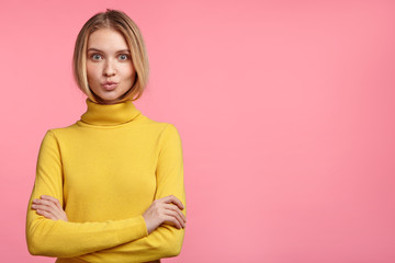 Beautiful female model rounds lips, keeps hands crossed, wears casual comfrotable sweater, poses against blank studio wall for your advertisment or hearder. Young woman advertises new outfit.