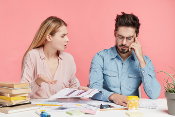 Indignant cute female worker tries to understand figures in financial report, looks at her male colleague, being angry with him, as he can`t explain anything, looks tired and exhausted of work