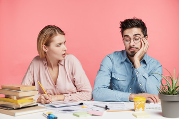 Naklejka premium Indignant woman writes notes, looks at lazy groupmate or colleague who doesn`t want work, prepare together scientific report, surrounded with many books, isolated over pink background. Team work