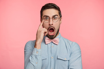 Portrait of handsome bearded man looks with shocked expression, keeps mouth opened, stares through spectacles, isolated over pink background. Amazed frightened male models in studio, hears bad news