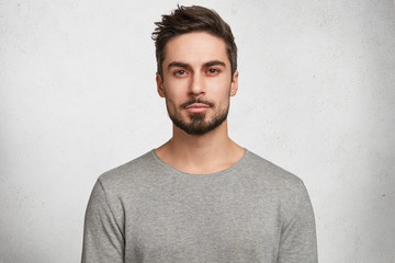 Isolated shot of young handsome male with beard, mustache and trendy hairdo, wears casual grey sweater, has serious expression as listens to interlocutor, poses in studio against white background - obrazy, fototapety, plakaty