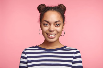 Portrait of cheerful dark skinned female with two hair buns, dressed in casual striped sweater, glad recieve good news about including in staff or puting on list, expresses positive emotions