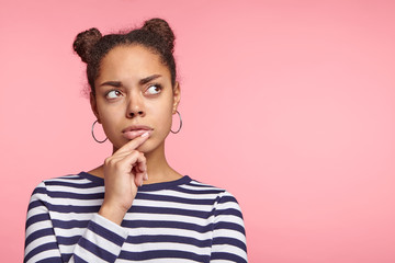 Mixed race female model looks pensively asise, tries to make decision, keeps finger on lips, poses agianst pink background with copy space for your promotional text. People and body language concept