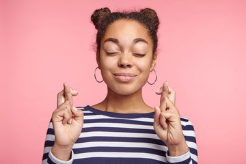 Isolated shot of attractive coquette female model with dark skin closes eyes and crosses fingers in anticipation, believes for good luck, isolated over pink studio background. Woman gestures indoors - obrazy, fototapety, plakaty