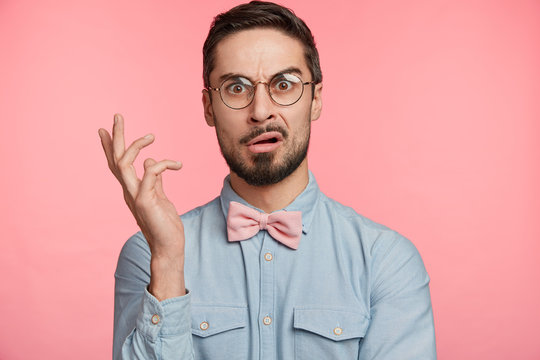 Portrait of dissatisdfied man wears round spectacles, has discontet, indigant look, gestures hand, being surprised with something, poses against pink studio background. Facial expression concept