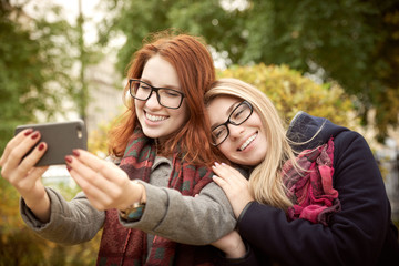 Fototapeta premium two young student girlfriend with long hair and glasses walking in the autumn Park dressed in a coat taking a selfie on the background of autumn Park in natural light