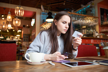 attractive stylish modern young woman who is shopping in the Internet sitting at a table in a cafe or bar with your smartphone and makes payment with a Bank card