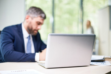 Fototapeta premium Busy open plan office: confident middle-aged manager wrapped up in work sitting at desk while his female colleague standing at panoramic window