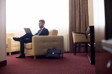 Full length portrait of handsome young businessman using laptop in hotel room, relaxing in armchair, copy space