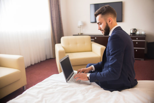 Side view portrait of handsome bearded businessman using laptop sitting on bed in comfortable hotel room during business travel, copy space