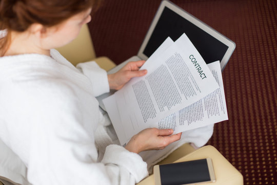 High angle portrait of young businesswoman dressed in bathrobe working with laptop and contract documents in hotel room during business travel, copy space