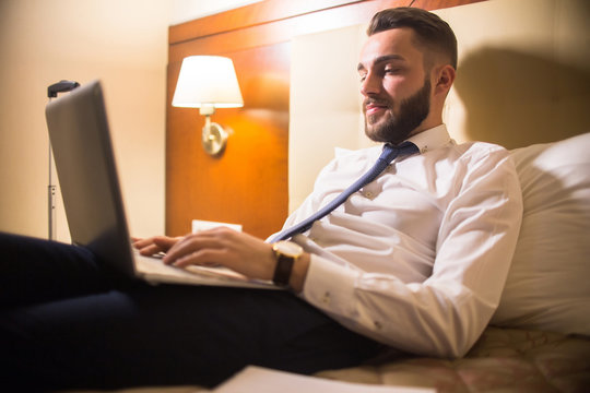 Portrait of handsome bearded businessman using laptop in bed enjoying hotel stay during business travel