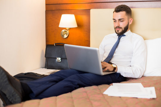 Full length portrait of handsome bearded businessman using laptop in bed enjoying hotel stay during business trip