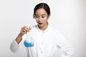 Fototapeta premium Pretty Chinese female laboratory assistant carrying out scientific research, studying chemical reaction, holding flask filled with blue solution. Chemistry, cosmetology, biology and science concept