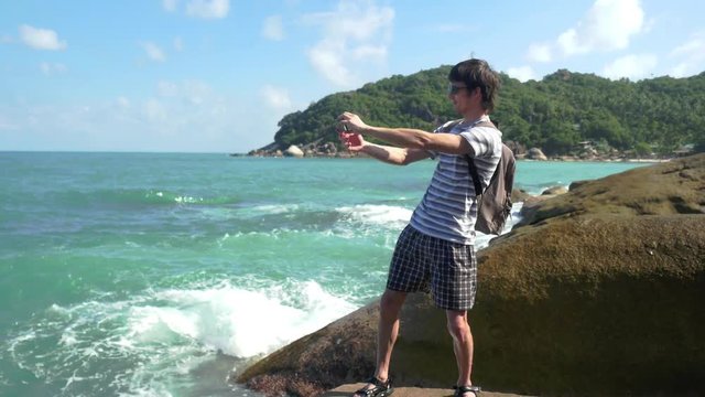 Happy man wearing sunglasses and backpack taking picture with smartphone on the rocky beach. Koh Samui. 3840x2160