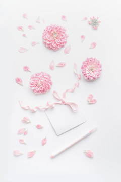 Pastel color mock up with pink flowers and petals, blank paper card with ribbon and point pen on white desktop background, top view. Layout of greeting card for Mothers day, wedding or happy event