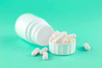 Close up white pill bottle with spilled out pills and capsules in cap on aquamarine background with copy space. Focus on foreground, soft bokeh. Pharmacy drugstore concept