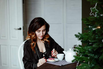 The young woman in a black silk pajamas has breakfast in kitchen in the apartment decorated by Christmas. The girl eats a tasty crimson dessert and has tea.