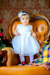 little girl in a beautiful dress is standing on a chair near a festive Christmas tree.