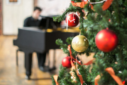 music, holidays, feast concept. close up of marvelous sparkling balls of golden and crimson colours, that hang on the christmas tree with red ribbons on the branches on the background with pianist