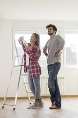 Young couple in empty room looking at the wall and think what color will be paint walls, she is leaning on ladder and holding tone card