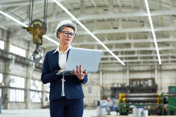 Pensive attractive businesswoman running industrial factory and visiting production workshop using portable computer while standing alone at construction site