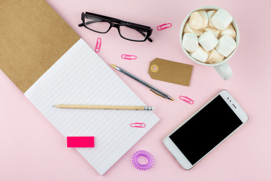 Creative flat lay photo of workspace desk with smartphone, eyeglasses, pen, pencil and notebook, minimal style on pink background. Minimal business concept with cocoa and marshmallow 
