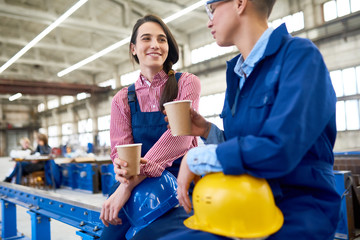 Positive young female construction workers drinking coffee from disposable cups and talking while resting during break