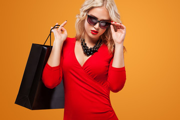 Blonde woman in sunglasses at shopping holding dark bag on orange background on black friday holiday. Copy space for sale ads.