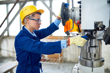 Serious confident female manual worker in hardhat wearing workwear adjusting drilling machine before operation