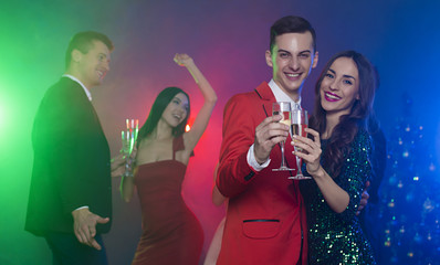 A group of young cheerful and happy friends are dancing at a party. A man and a woman are dancing in the foreground and clinking glasses with champagne