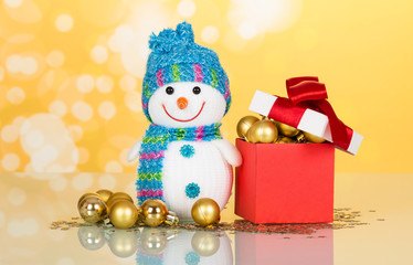 Charming little snowman in a hat next box filled with toys, balls, Christmas decorations on yellow