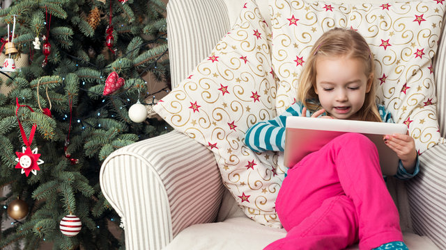 Cute preschool girl writing letter to santa on digital tablet, sitting in front of xmas tree