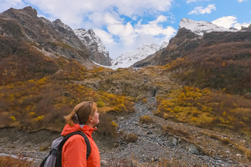 Fototapeta premium Landscape in the autumn Caucasian mountains with a woman tourist looking up at snowy peaks and autumn slopes