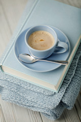 Coffee in a light blue cup and book in blue binding with a knitted scarf on a wooden background.