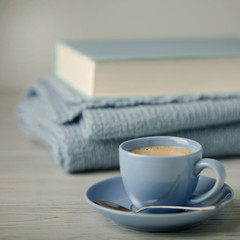Coffee in a light blue cup and book in blue binding with a knitted scarf on a wooden background.