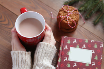 Female hands in the white sweater hold a red mug of cocoa on the wooden table