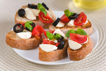 Bruschetta italian snack sandwiches with mozzarella, tomatoes and black olives decorated by basil leaves.