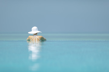 Fototapeta na wymiar Young woman in a white hat lounging in an infinity pool looking out to the ocean view in paradise