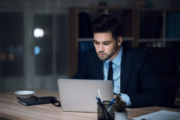 A man is sitting at a laptop in a dark office. The man in the suit works.