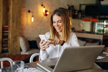 Young business woman reading message on a smartphone with a laptop in front of her