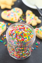 Fototapeta na wymiar Colorful sugar sprinkles in glass jar with decorated sugar cookies and icing in a bowl in the back, photographed on slate (Selective Focus, Focus one third into the sprinkles)