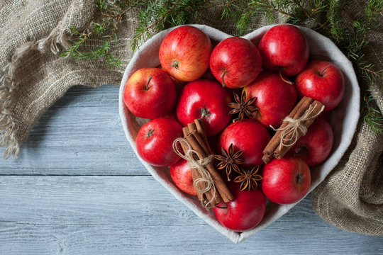 Red apples, basket in the form of heart on a wooden background, cinnamon, anise and branches of a juniper.